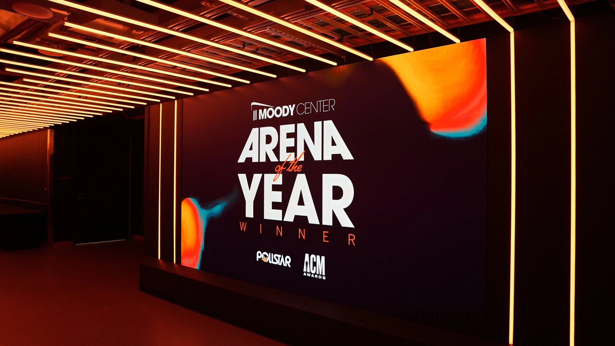 A large indoor digital screen displaying the text "Moody Center Arena of the Year Winner" with the logos for Politstar and ACM Awards at the bottom. The screen is framed by orange strip lights on a dark wall with a ceiling decorated with orange and white linear lights.