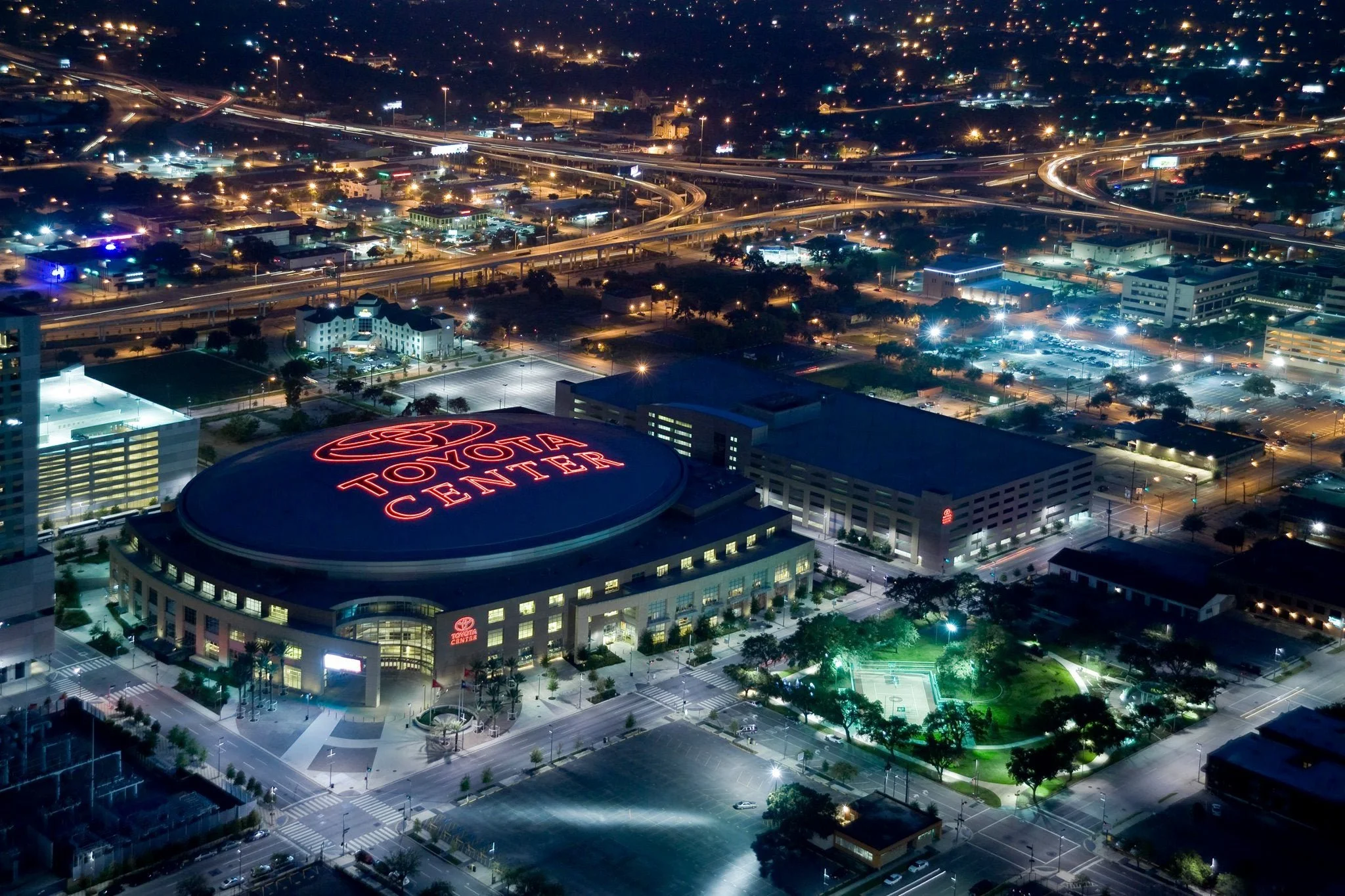 Night aerial view of Toyota Center with surrounding city lights, roads, parking lots, and a lit parking lot sports court.
