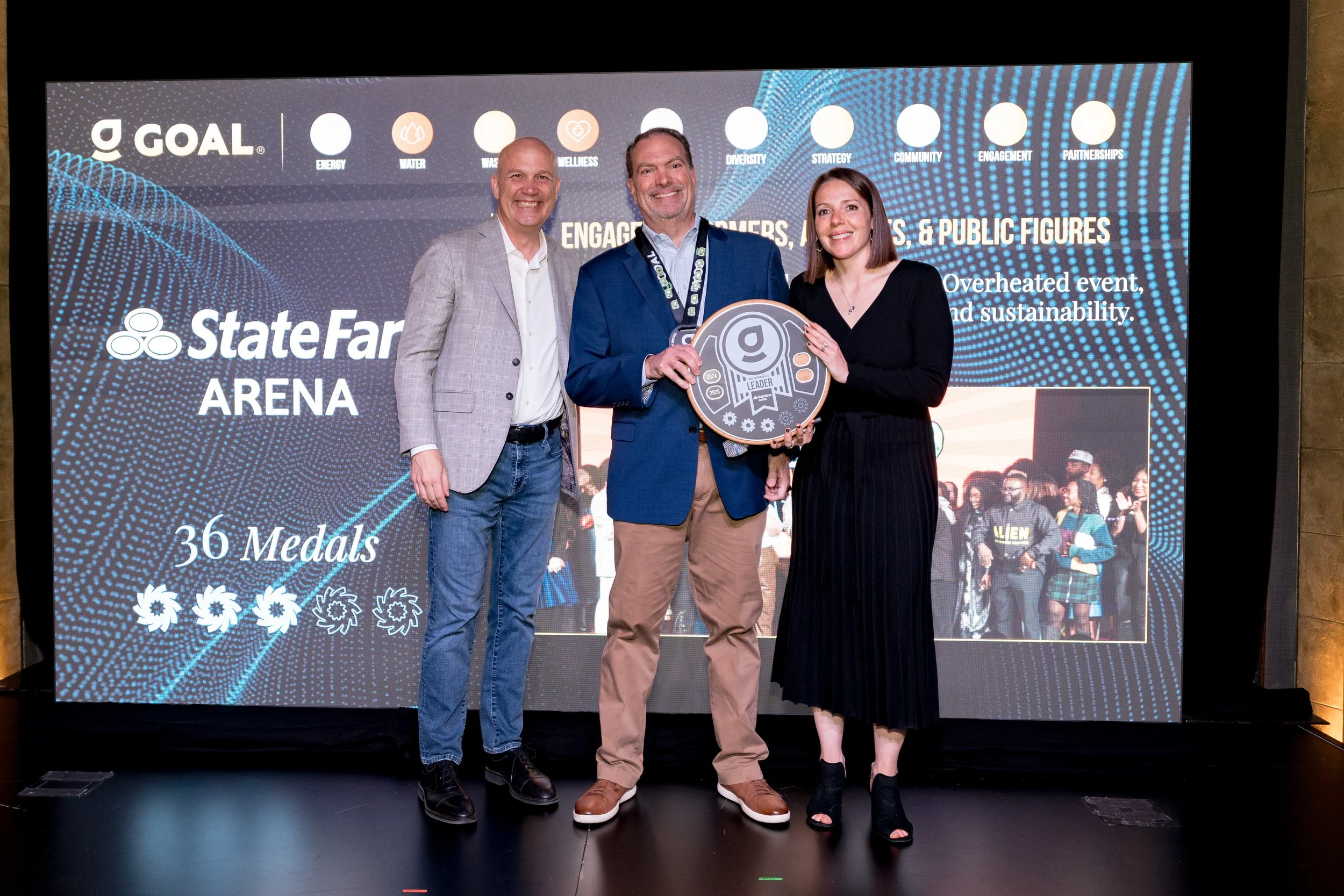 Three people are standing on a stage, smiling and holding a large award plaque, with a large screen behind them displaying the event details, including 'StateFarm Arena', '36 Medals', and the GOAL logo.
