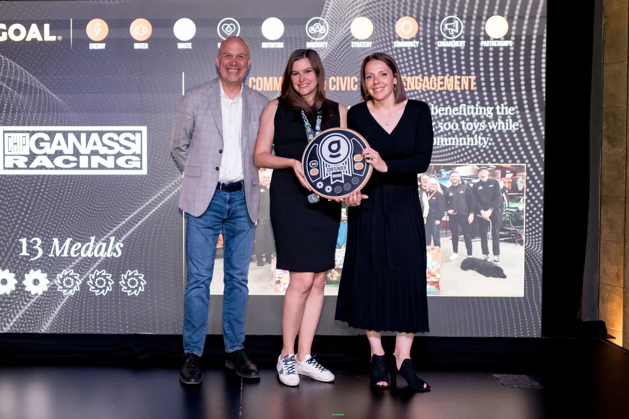 Three people standing on a stage, one woman holding an award, in front of a screen displaying the GOAL and Gannass Racing logos, with text about medals and community engagement.