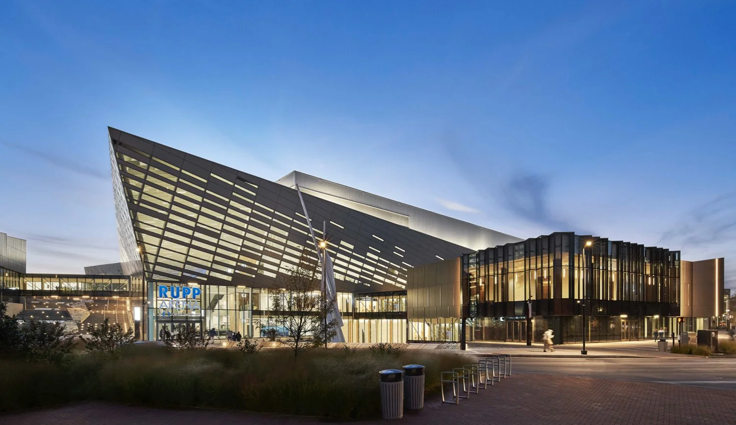 Exterior view of a modern sports arena building with illuminated signage reading RUPP ARENA, featuring sharp architectural angles and glass sections, taken during dusk.