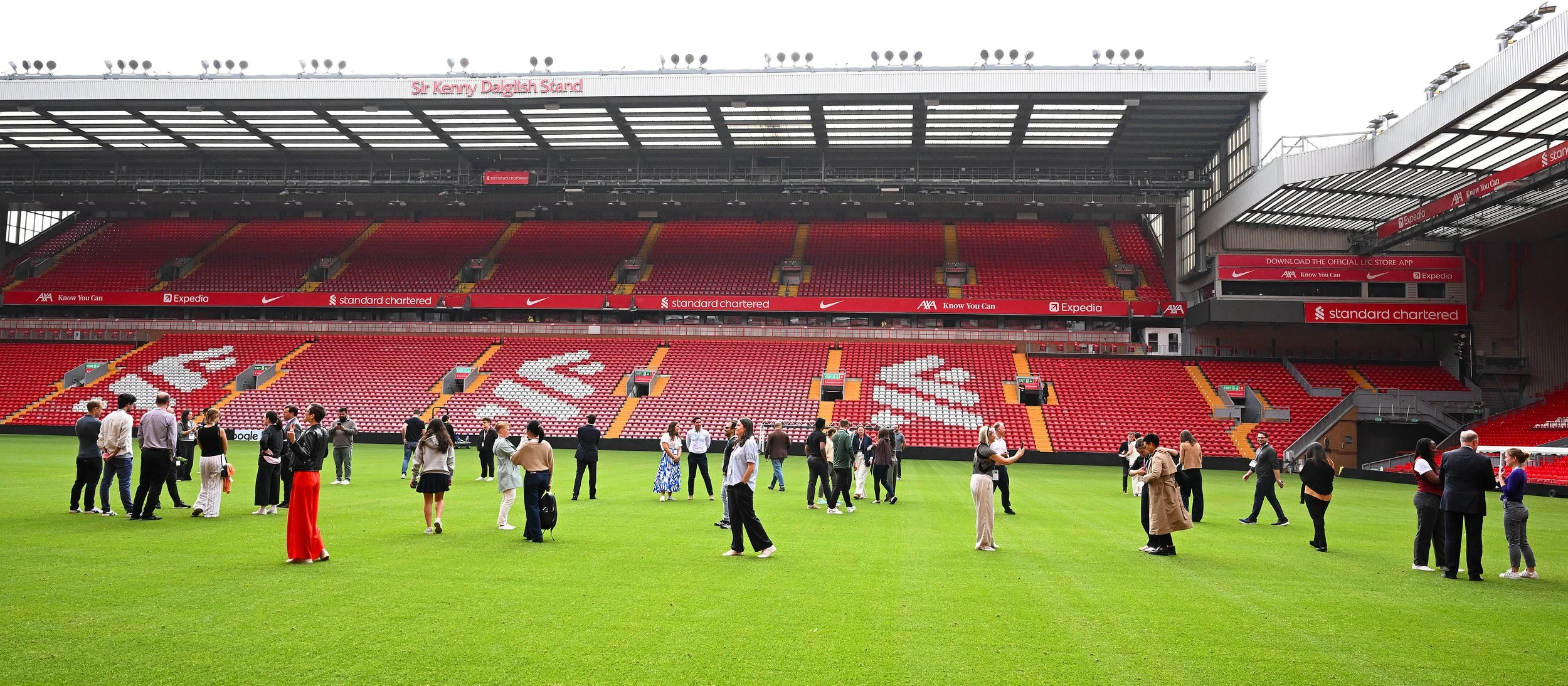 People standing and walking on the grass field inside a large empty sports stadium with red seats, advertising banners, and a roof structure overhead.
