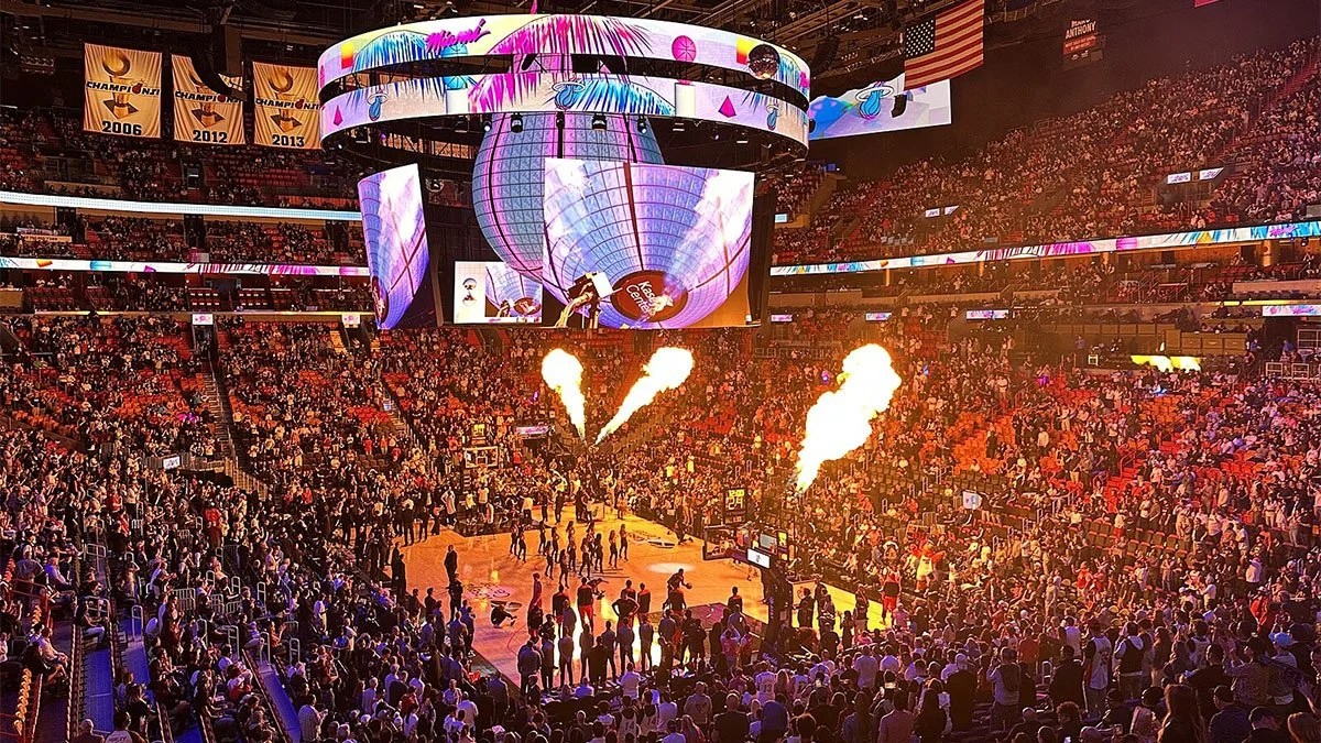 A packed basketball arena with a crowd, large digital screens displaying colorful graphics, pyrotechnics exploding on the court, and banners hanging from the rafters.