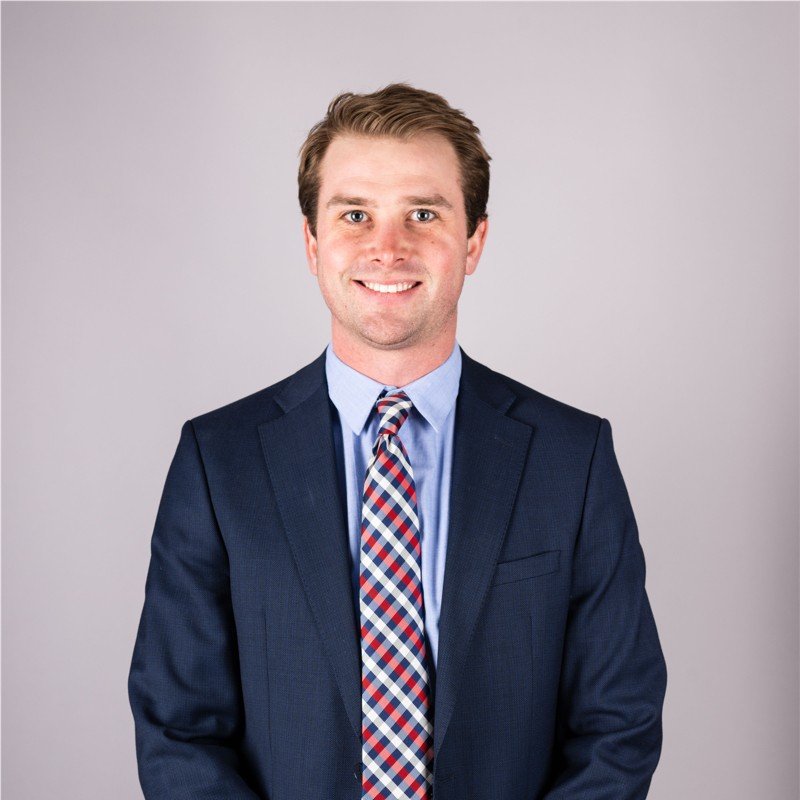 A young man in a navy suit, light blue shirt, and a colorful striped tie, smiling against a plain gray background.
