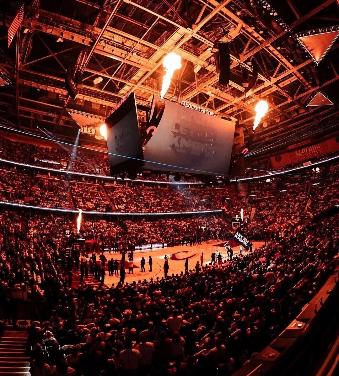 An indoor basketball arena filled with spectators, with bright orange lighting and pyrotechnics on the ceiling, a large electronic scoreboard hanging above the court.