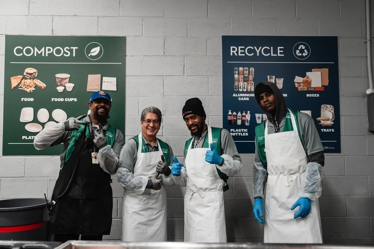 Four people in aprons and gloves standing in front of compost and recycle signs, smiling and giving thumbs up inside a facility.