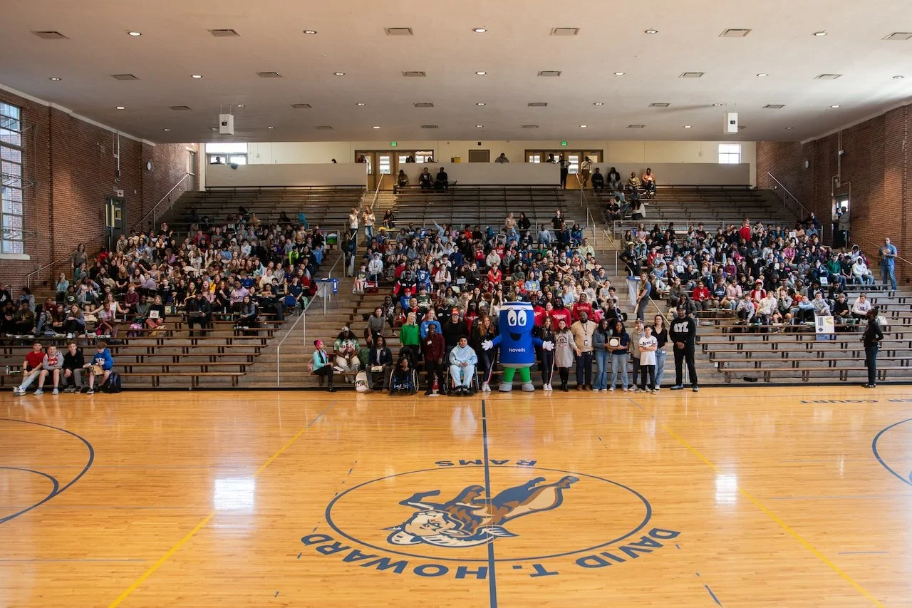 Large indoor gymnasium with wooden basketball court and a central logo, surrounded by many seated spectators and some standing. A person in a blue mascot costume and several people pose for a photo at the front. The gym has brick walls, large windows, and stairs leading to an upper level.