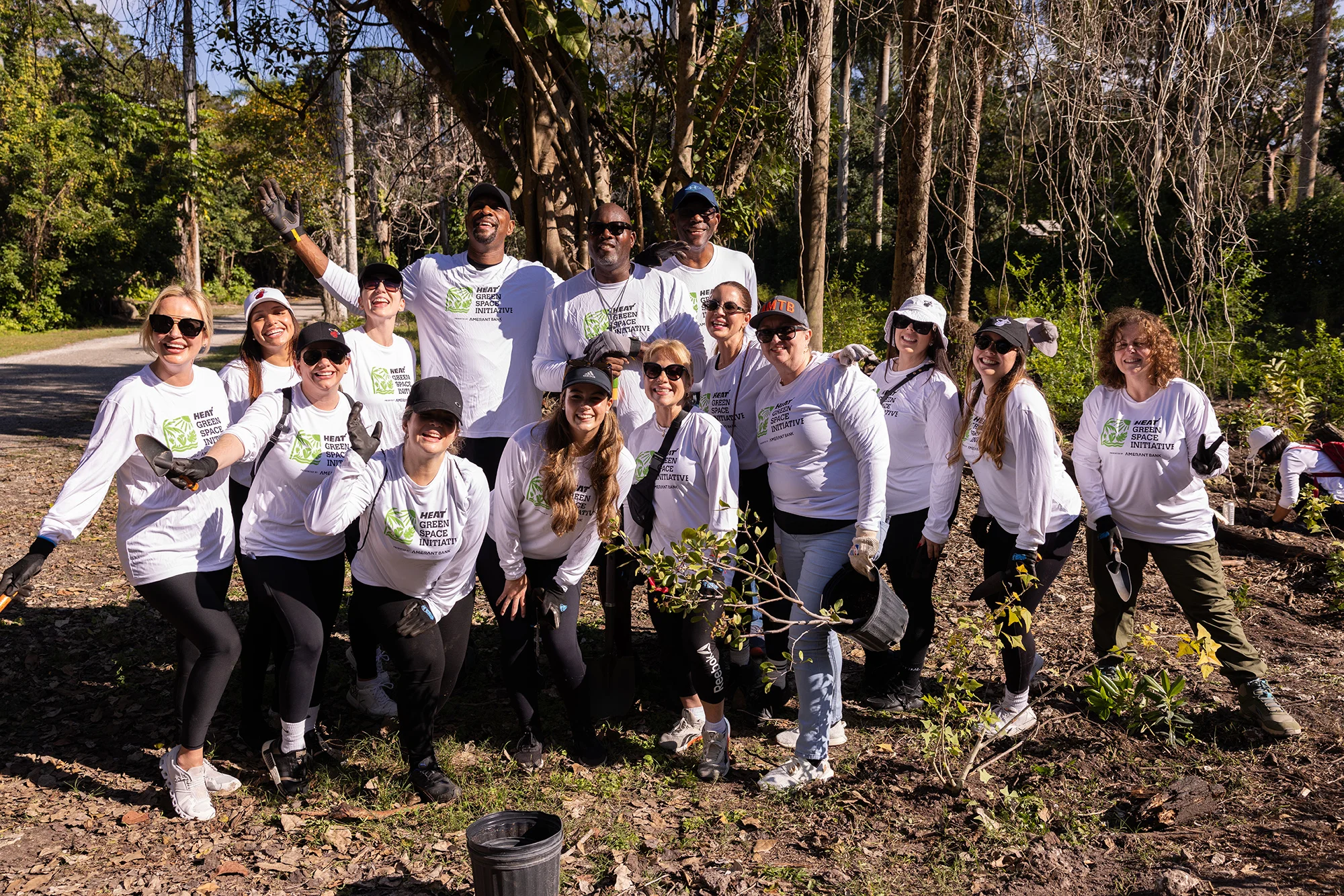 Group of volunteers wearing white shirts with the 'Heat Green Space Initiative' logo, planting trees outdoors in a wooded area.