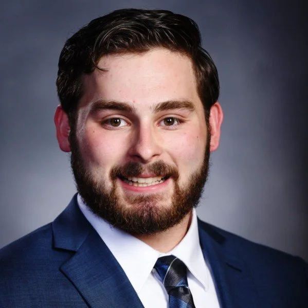 A young man with dark hair and a beard smiling, dressed in a navy suit, white shirt, and patterned tie, against a dark gradient background.