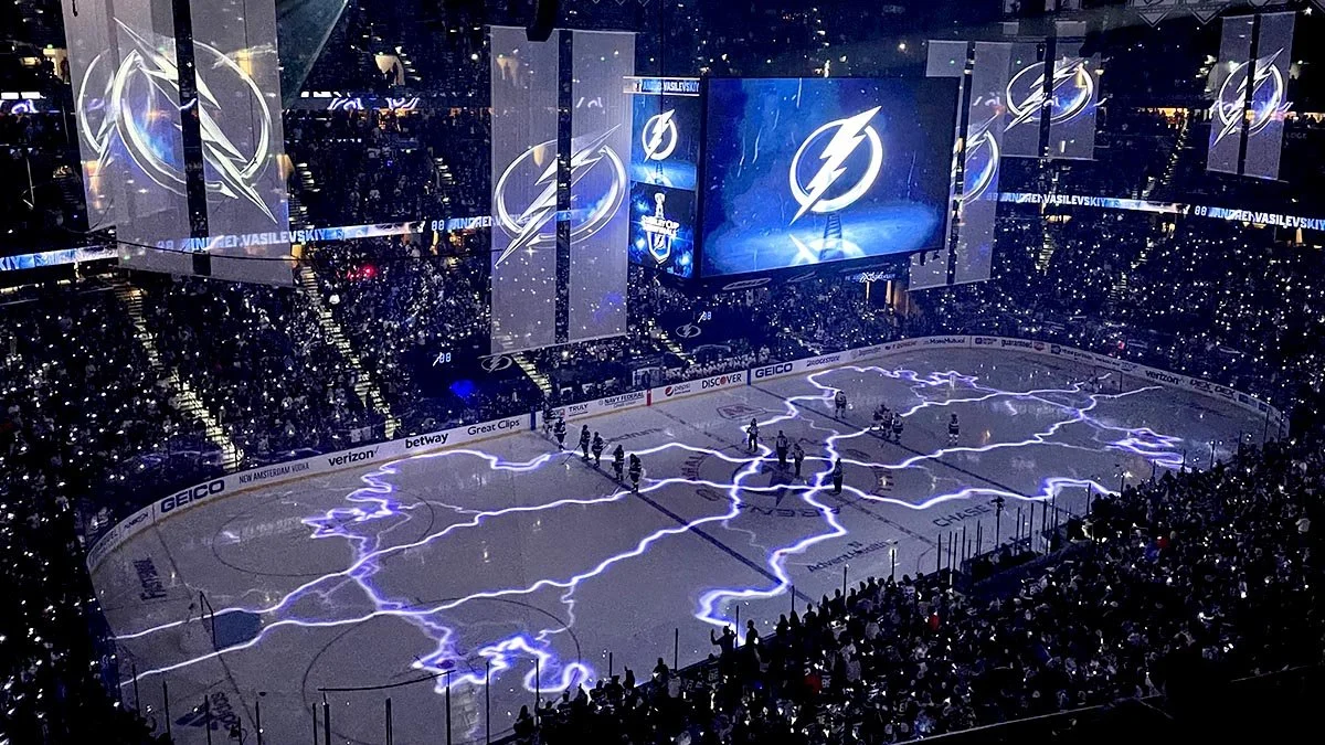 An ice hockey arena filled with spectators, with the Tampa Bay Lightning logo projected onto the ice and hanging screens. The ice surface features a lightning bolt design, and the arena is lit with blue lighting.