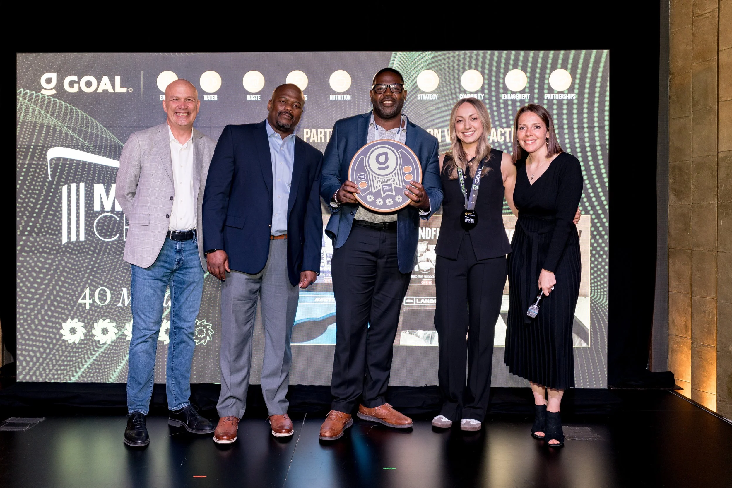 Group of five people on stage at an awards ceremony, with a large screen behind them displaying logos and text. The man in the middle holds a circular award, smiling at the camera.