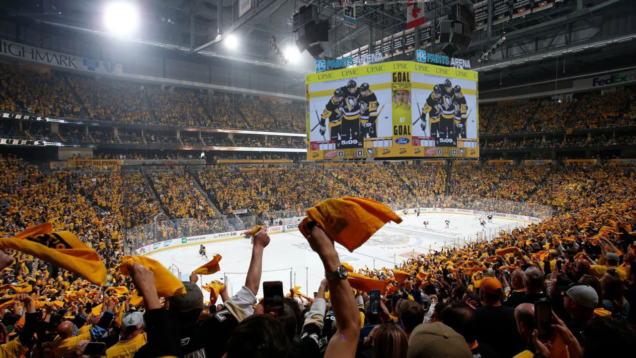 Crowd of hockey fans inside an arena, watching a game, waving yellow towels, with a large scoreboard showing players and a goal.