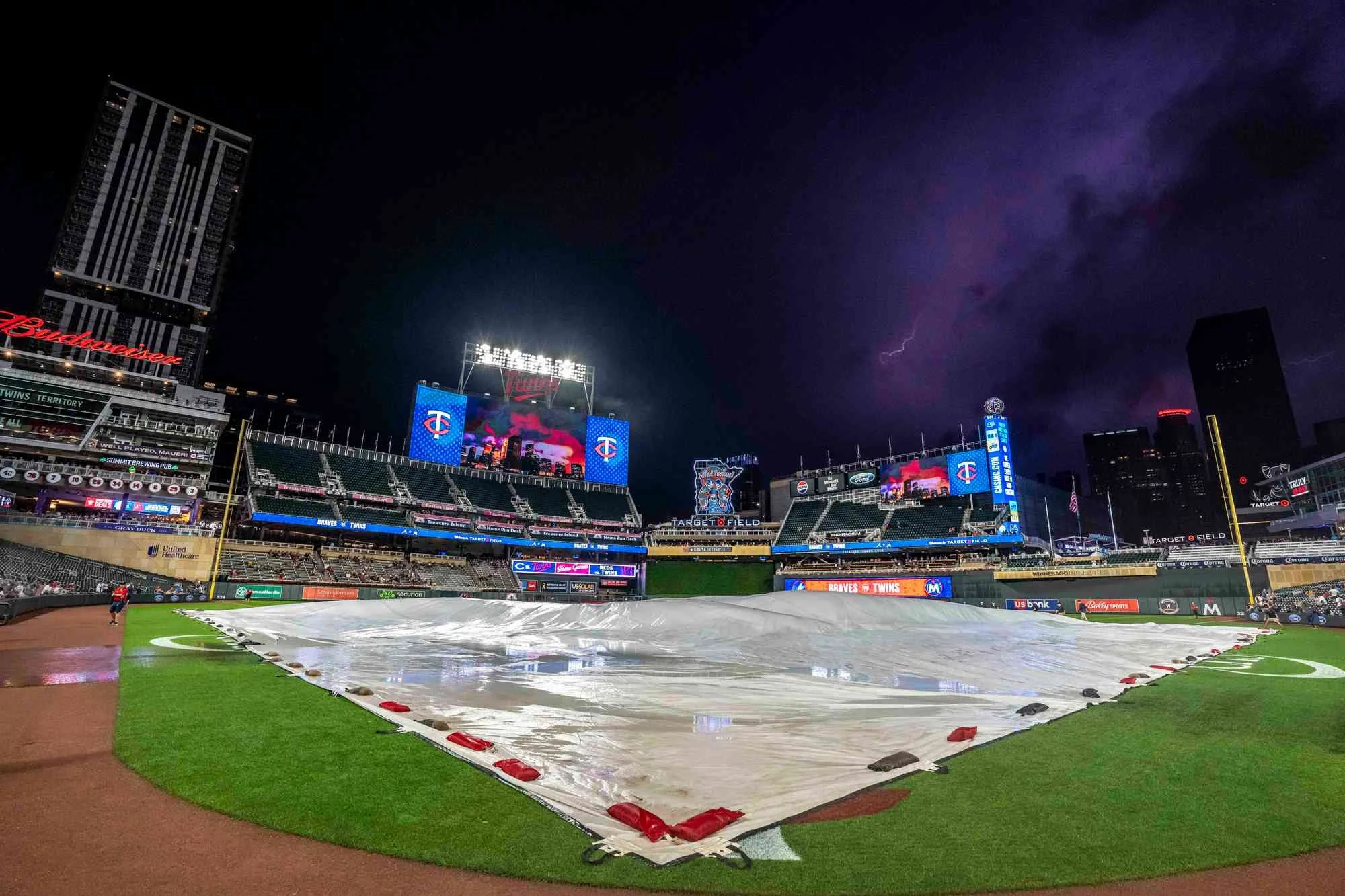 Empty baseball stadium at night with a tarp covering the field, lightning in the dark sky, and digital displays showing the Minnesota Twins logo.