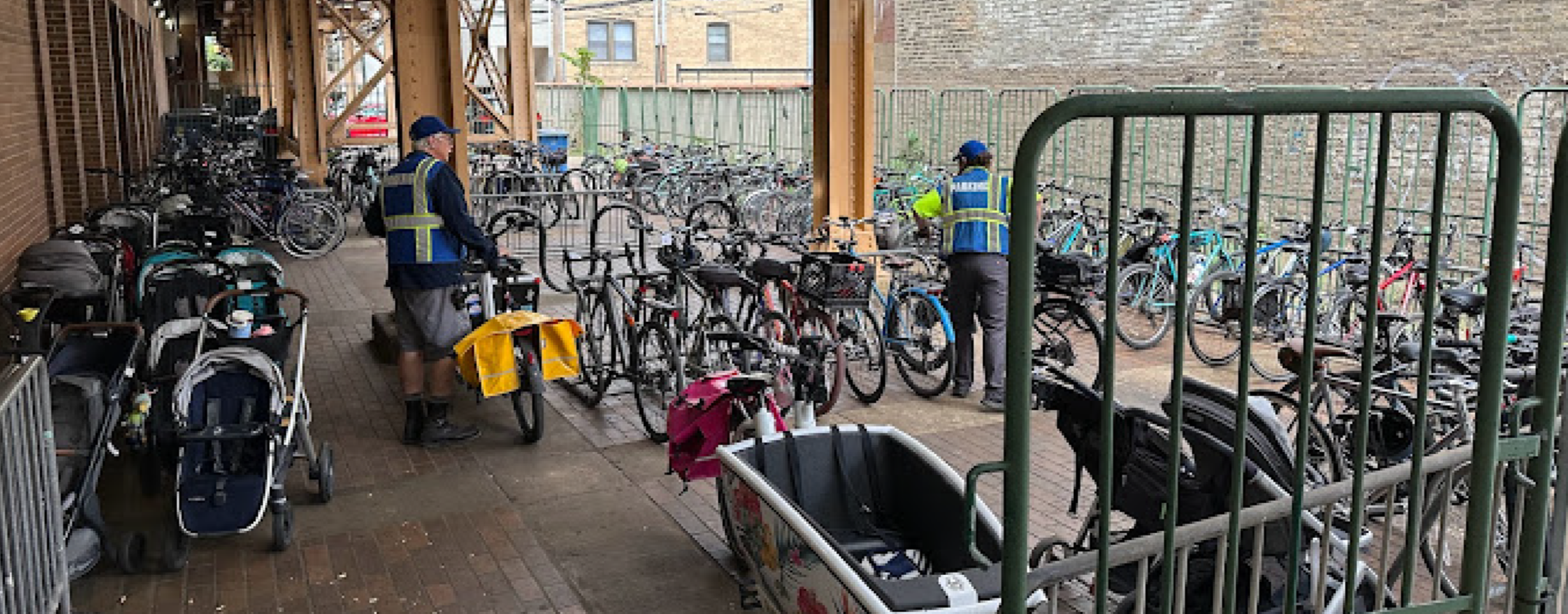 Several bicycles parked in a bike rack, with two police officers in high-visibility vests inspecting the area, in an outdoor urban space with brick walls and a wooden structure.