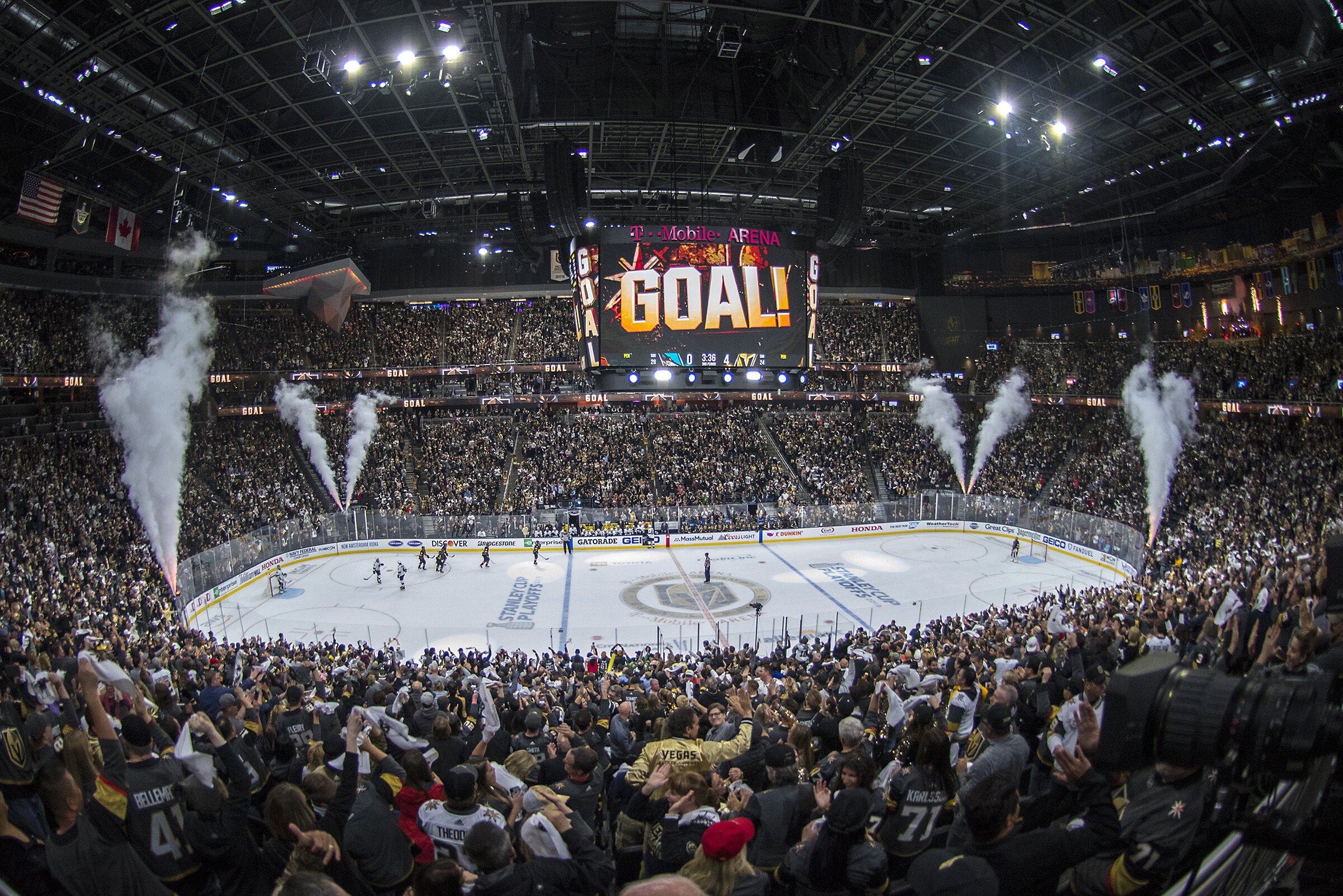 An indoor ice hockey arena filled with fans, with a large LED scoreboard displaying the word 'GOAL!' and a countdown timer, and players on the ice celebrating.
