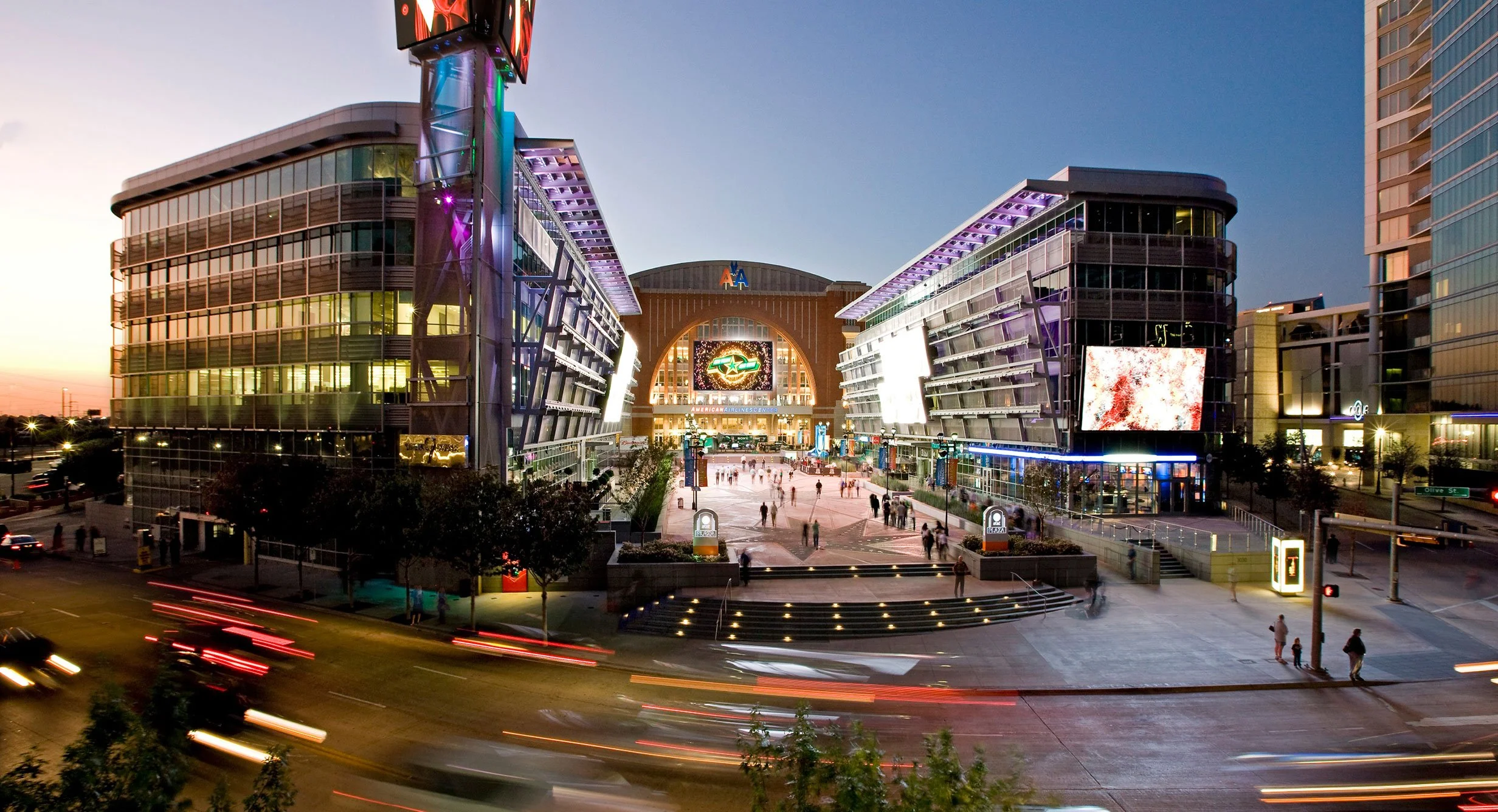 Night view of a modern shopping center with illuminated shops, people walking, and blurred vehicle lights on the street in front.