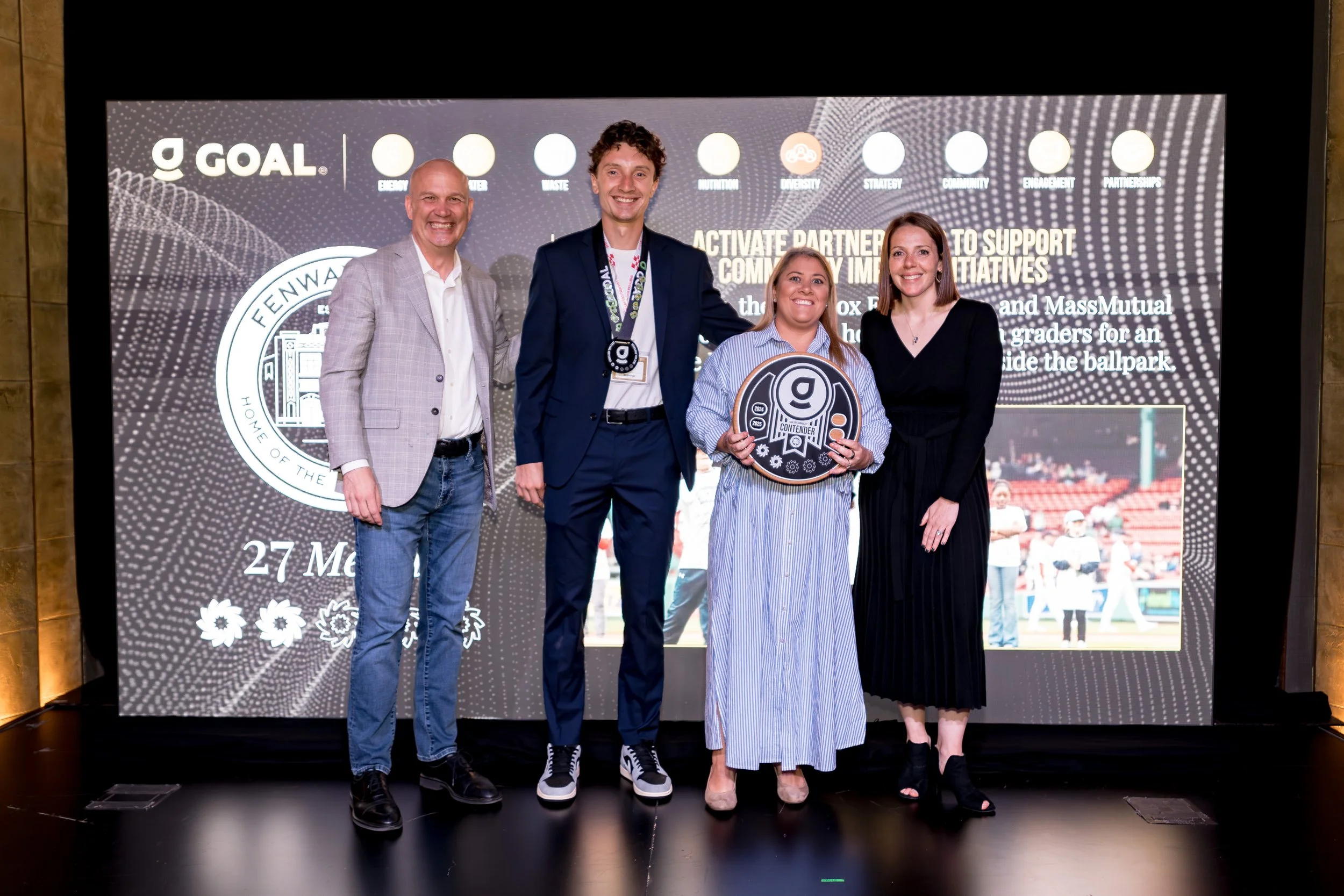 Four people standing on a stage in front of a large digital screen, with three men and one woman holding an award, celebrating at an event. The screen behind them features logos and text related to community initiatives and a park event.