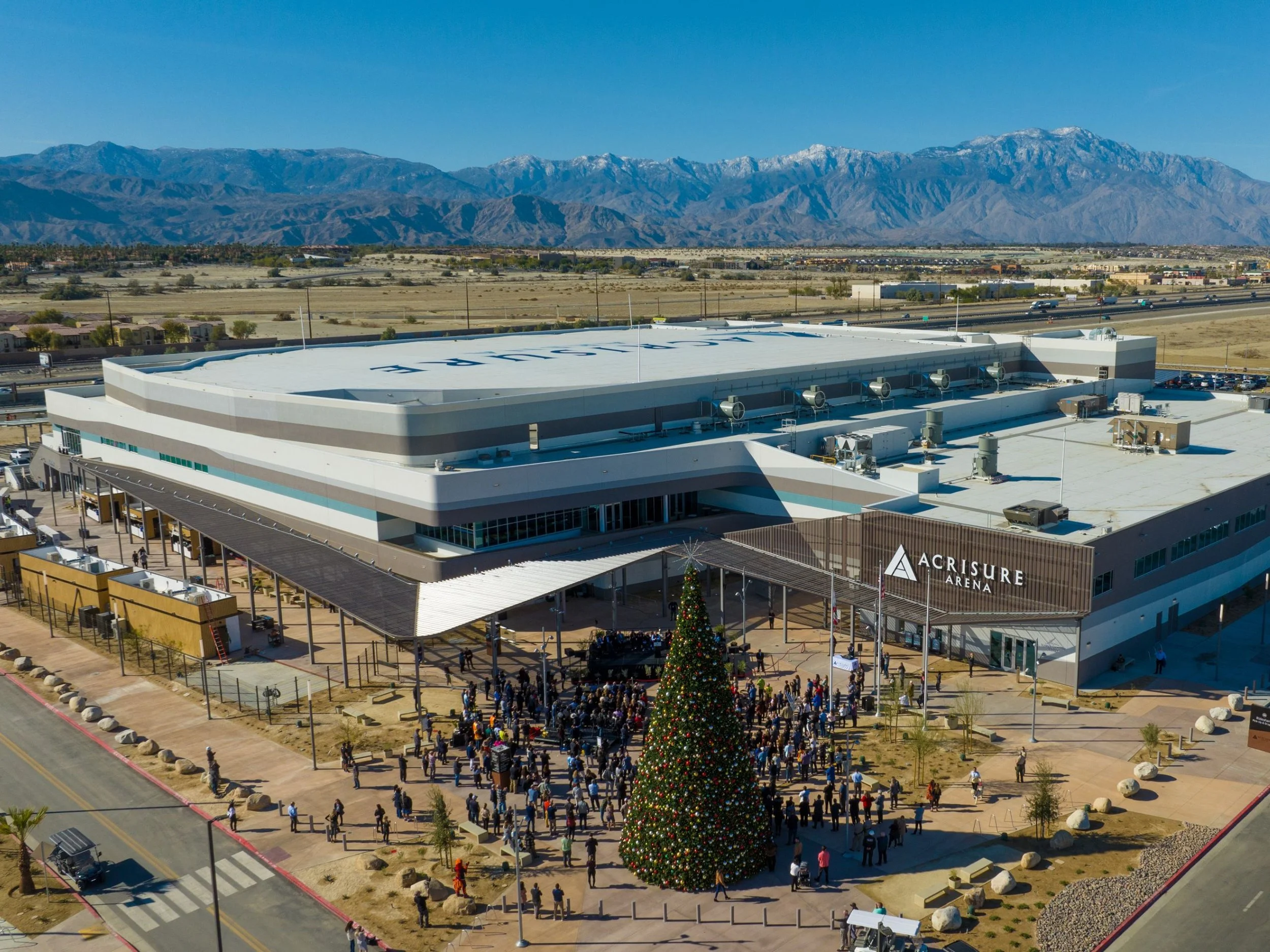 A large arena with a Christmas tree and crowds outside on a sunny day, with mountains in the background.