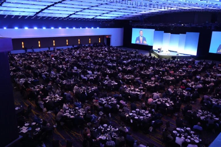 Large conference hall filled with attendees seated at tables, watching a speaker on stage and a large screen displaying the speaker's image.