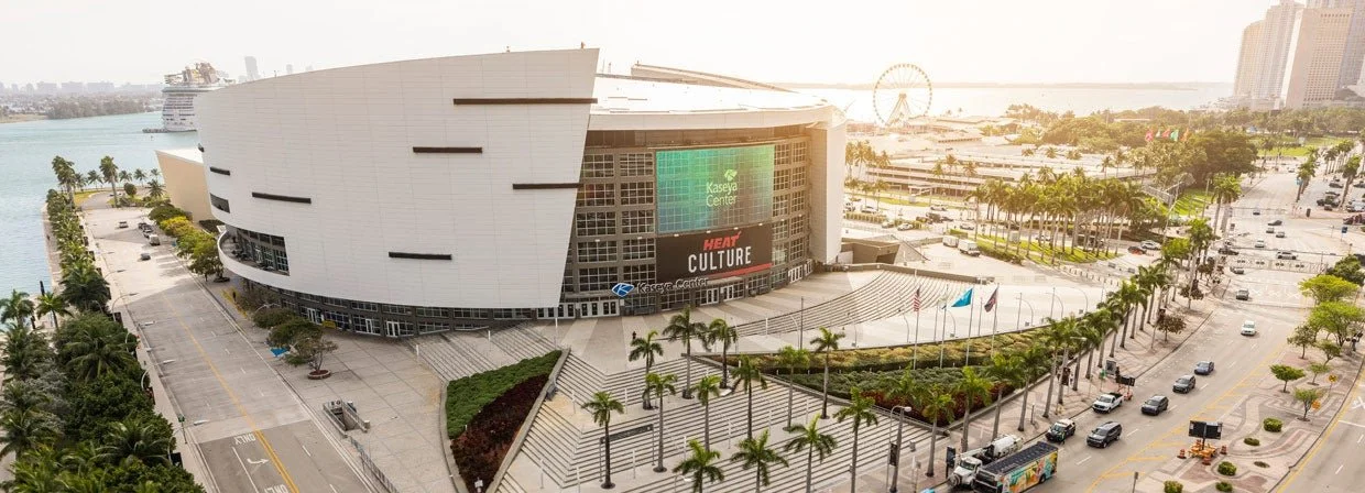 Aerial view of the Kaseya Center in Miami, Florida, with palm trees, streets filled with cars, and a distant Ferris wheel.