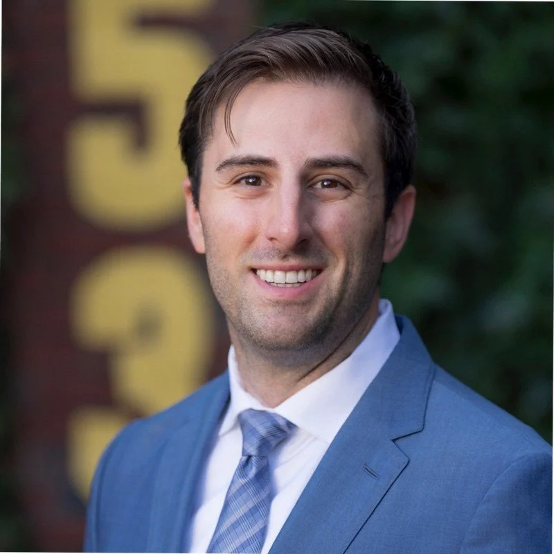 A man in a blue suit and tie smiling outdoors with blurred background.