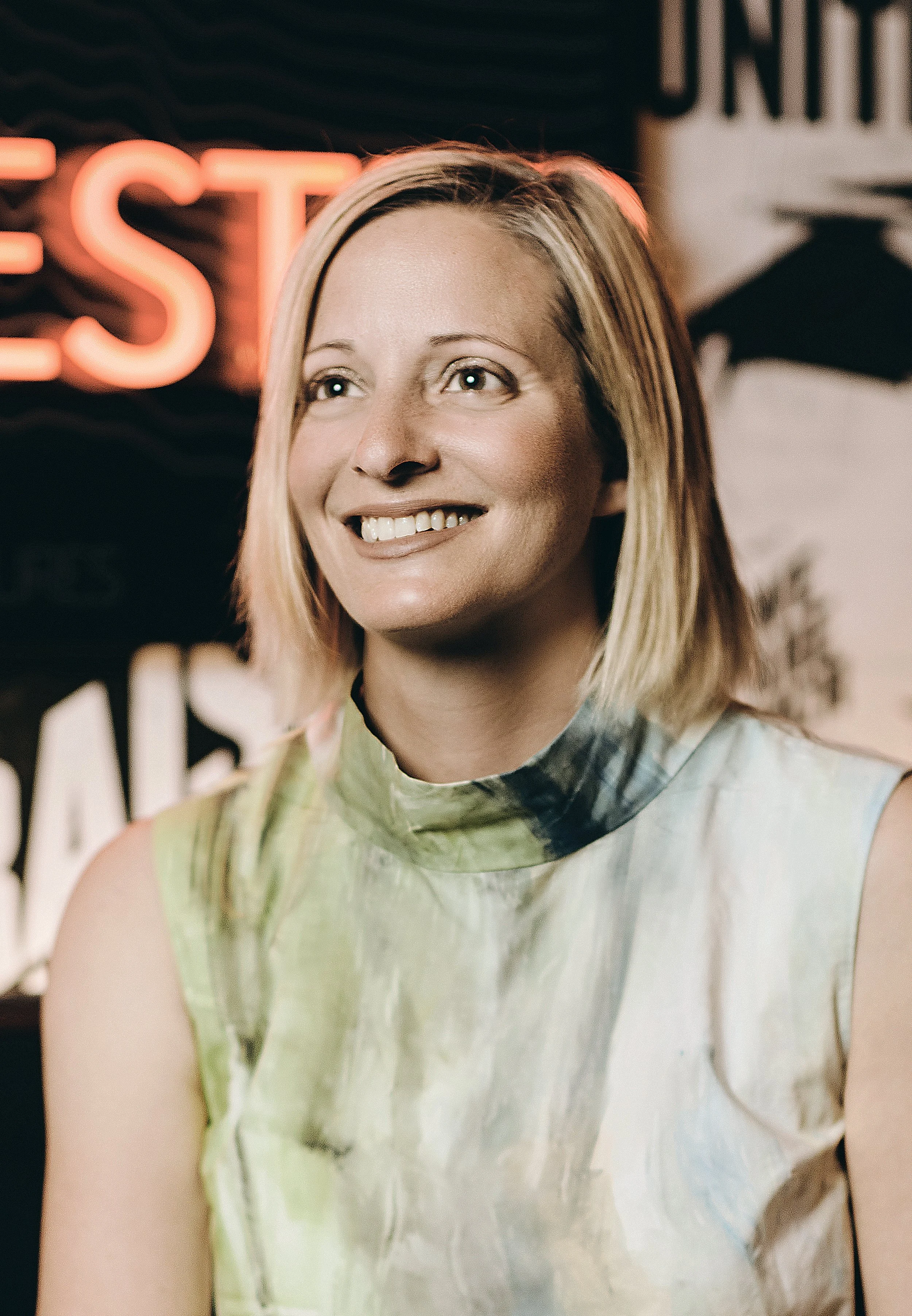 A woman with shoulder-length blonde hair smiling indoors, with a neon sign in the background.