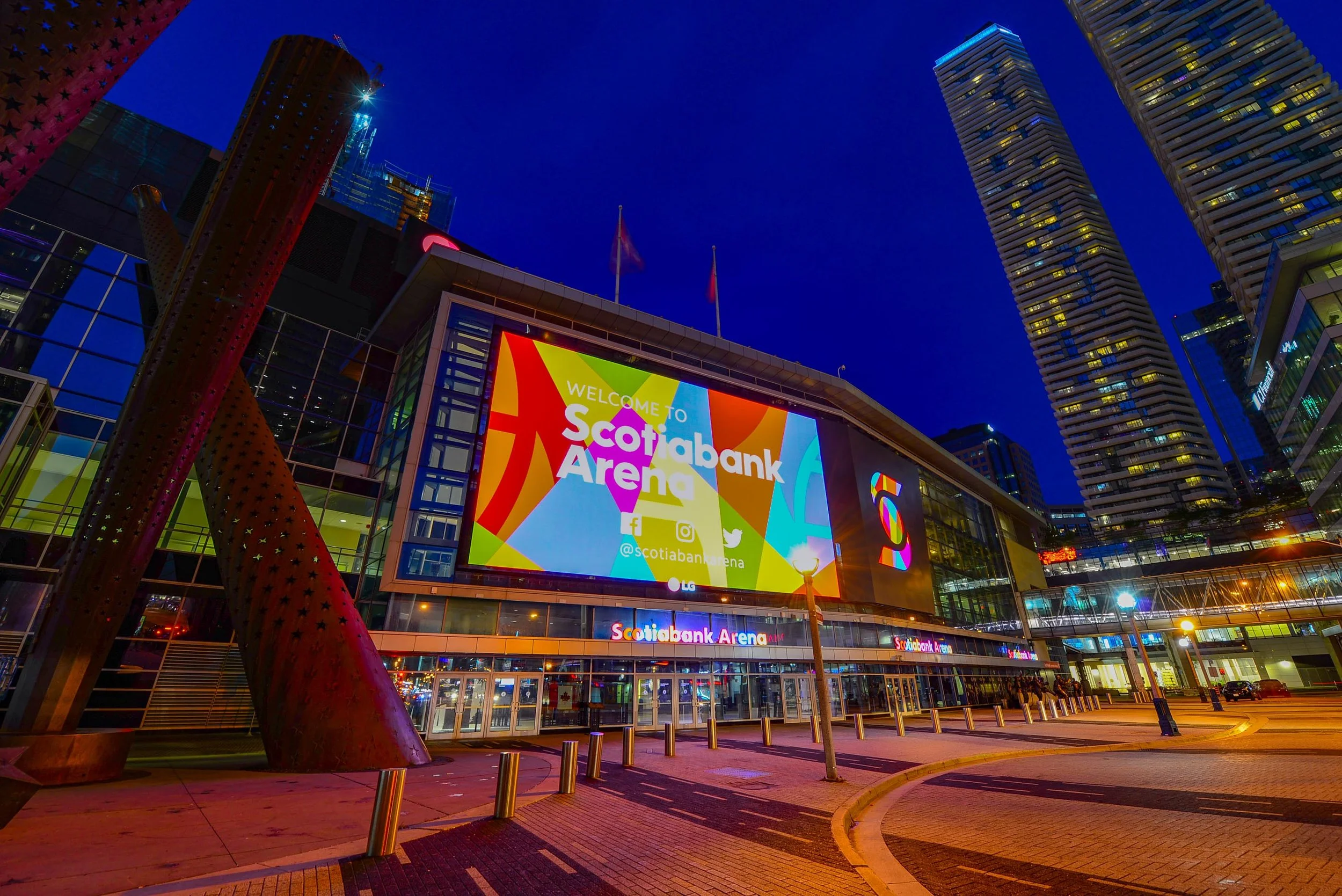 Night view of Scotiabank Arena in Toronto, Canada, with large colorful digital billboard displaying welcoming message, surrounding modern buildings, and streetlights illuminating the sidewalk.