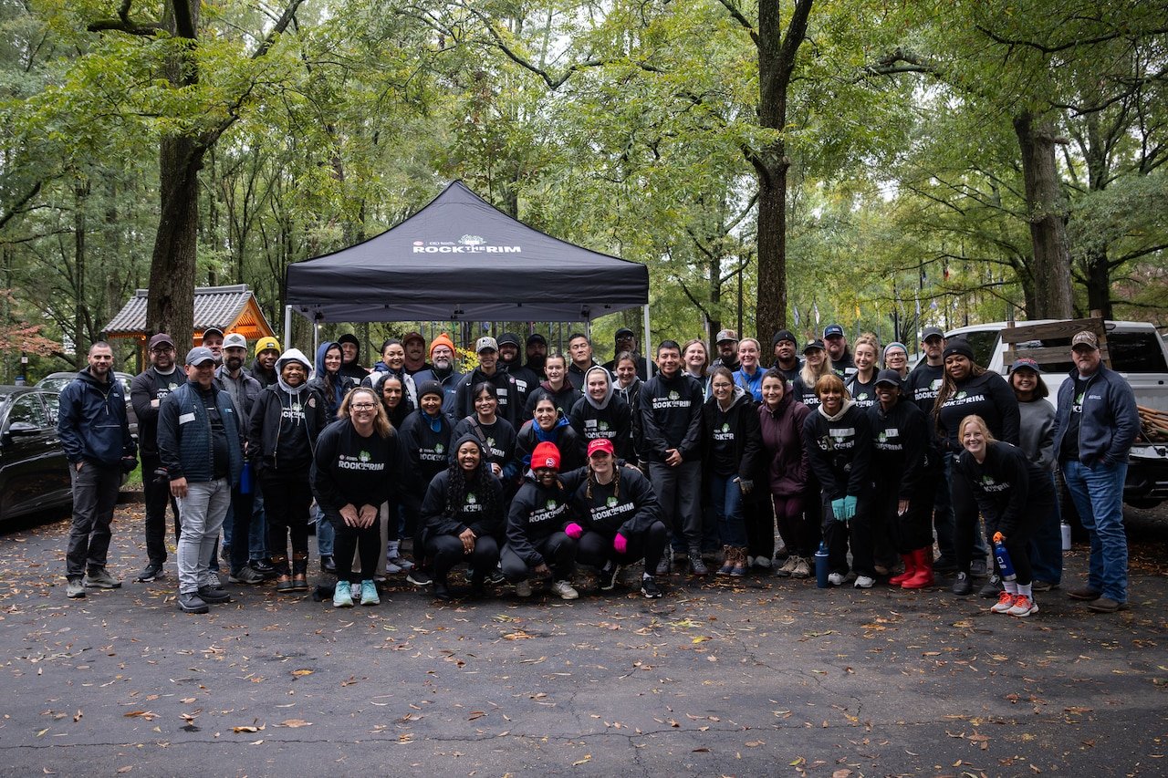 Group of people gathered outdoors in fall, posing for a photo in front of a black canopy with a wooded background. They are dressed warmly, some wearing jackets and hats.
