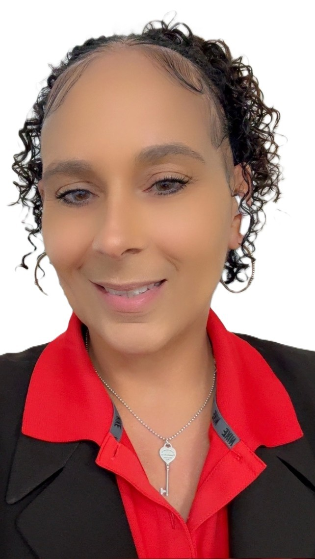 A woman with curly hair and light makeup smiling, wearing a black blazer over a red collared shirt, accessorized with a necklace featuring a key pendant.