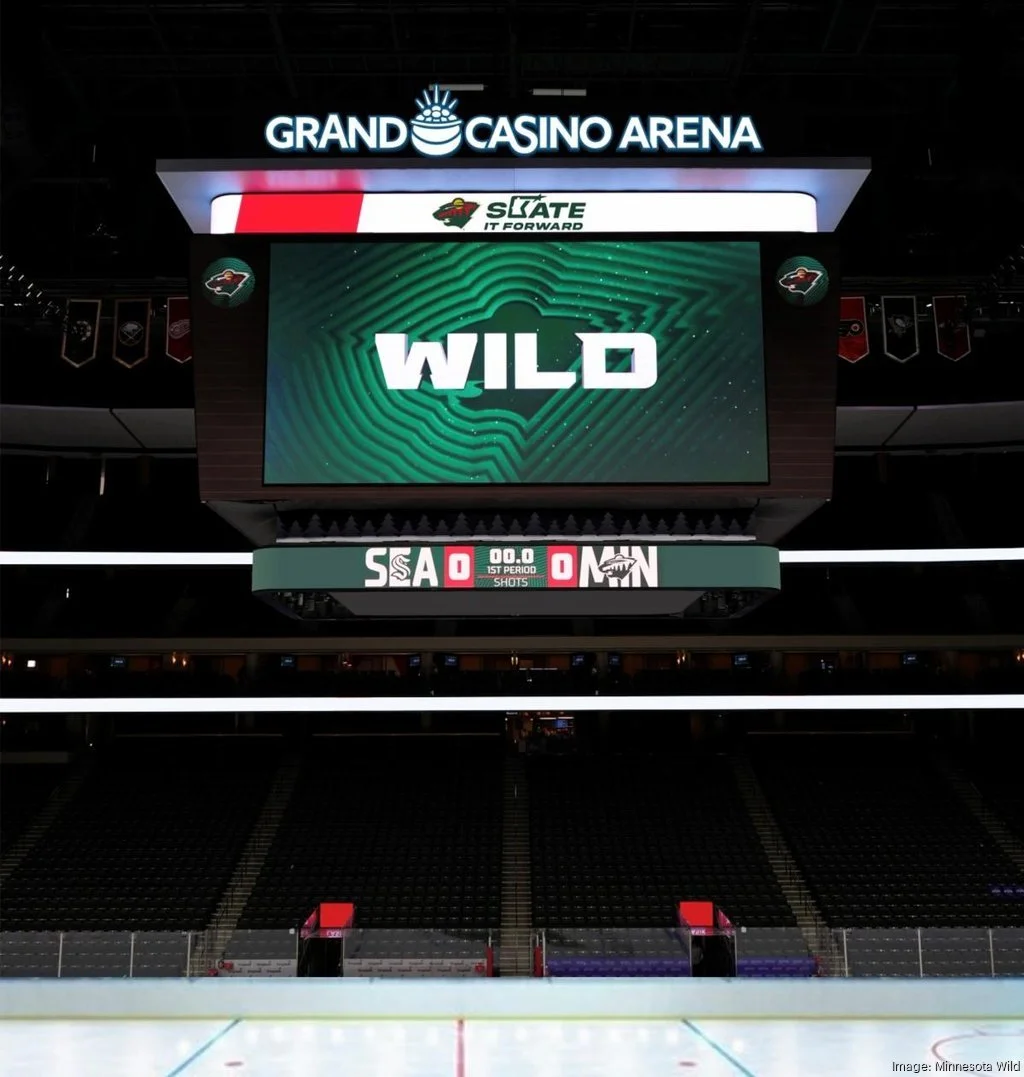 Empty hockey arena with a large scoreboard displaying the word "WILD" and the logo of the Minnesota Wild. The arena is dark, and the lower section of the ice rink is visible.
