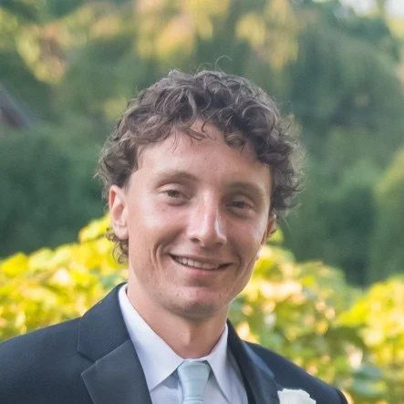 A young man with curly hair wearing a dark suit, light blue tie, and white shirt, smiling outdoors with yellow flowers and green trees in the background.