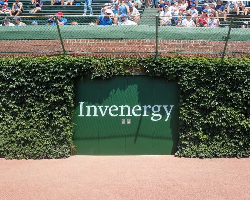 A green wall with the word 'Invernergy' in white lettering, surrounded by ivy, and a green door below the sign, with spectators in the background at a sports event.