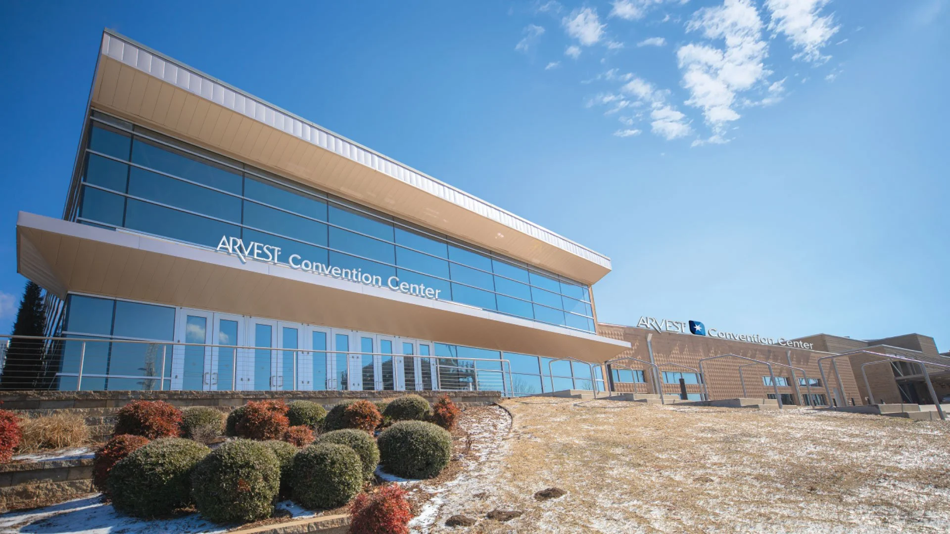 Exterior of the Arvest Convention Center with a clear blue sky, bushes, and a snowy ground.