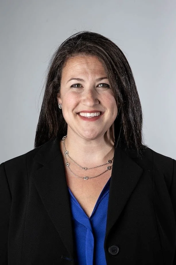 A woman with shoulder-length dark hair, wearing a black blazer and a blue blouse, smiling at the camera against a plain gray background.