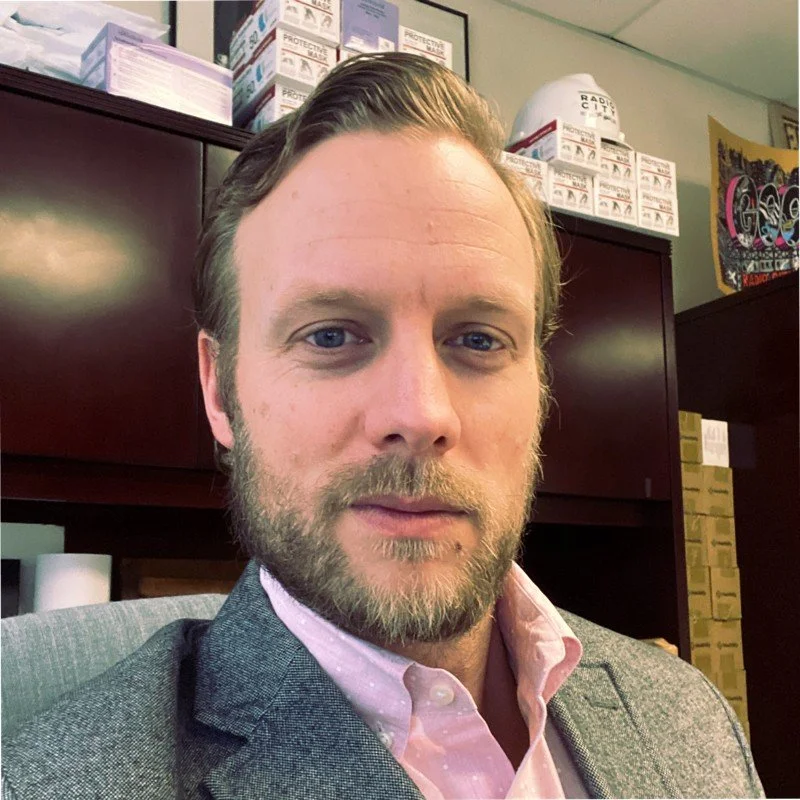 A man with a beard and light brown hair, wearing a pink dress shirt and a gray blazer, sitting in an office or retail environment with shelves of boxes and items behind him.