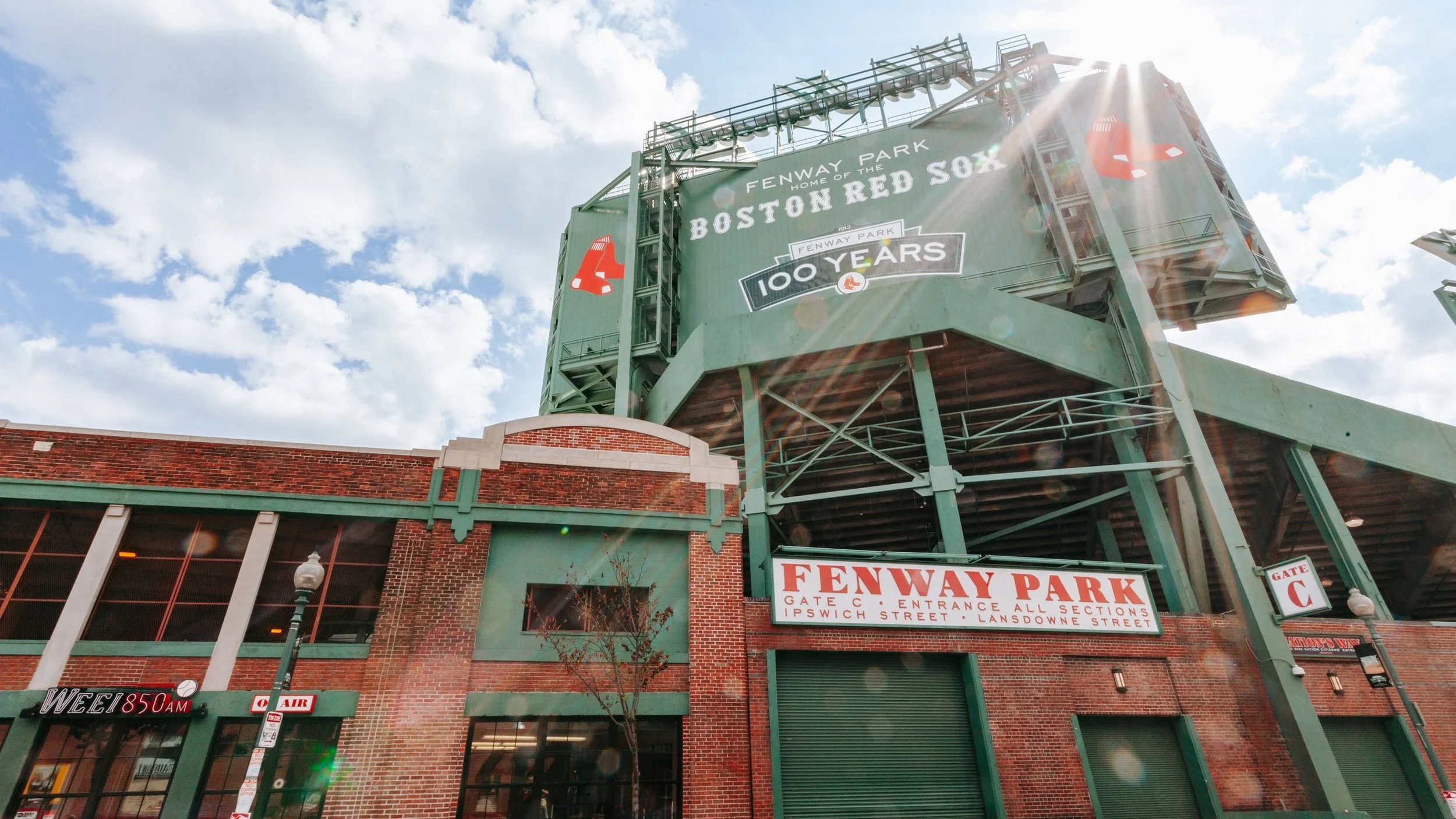 Exterior view of Fenway Park, home of the Boston Red Sox, with a large green sign celebrating its 100th anniversary and the entrance labeled Gate C, under a partly cloudy sky with sunlight lens flare.