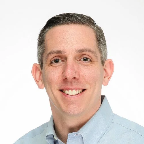 Headshot of a man with short gray hair, wearing a light blue collared shirt, smiling against a white background.