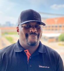 A man wearing a black cap and polo shirt standing outdoors.