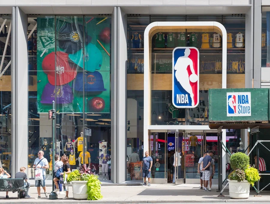 Exterior of an NBA Store with large glass windows displaying merchandise such as jerseys, hats, and basketballs, and an NBA logo sign above the entrance.
