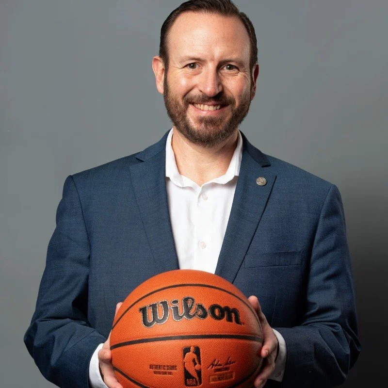 A man in a blue suit holding a Wilson NBA basketball, smiling at the camera.