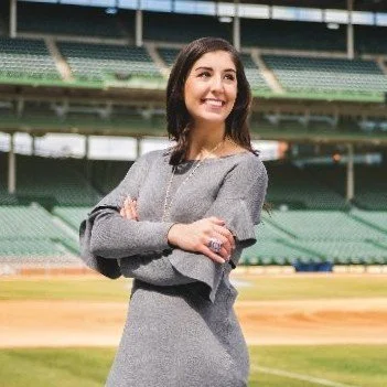 A young woman standing on a baseball field, smiling and looking to the side, wearing a gray dress with her arms crossed.
