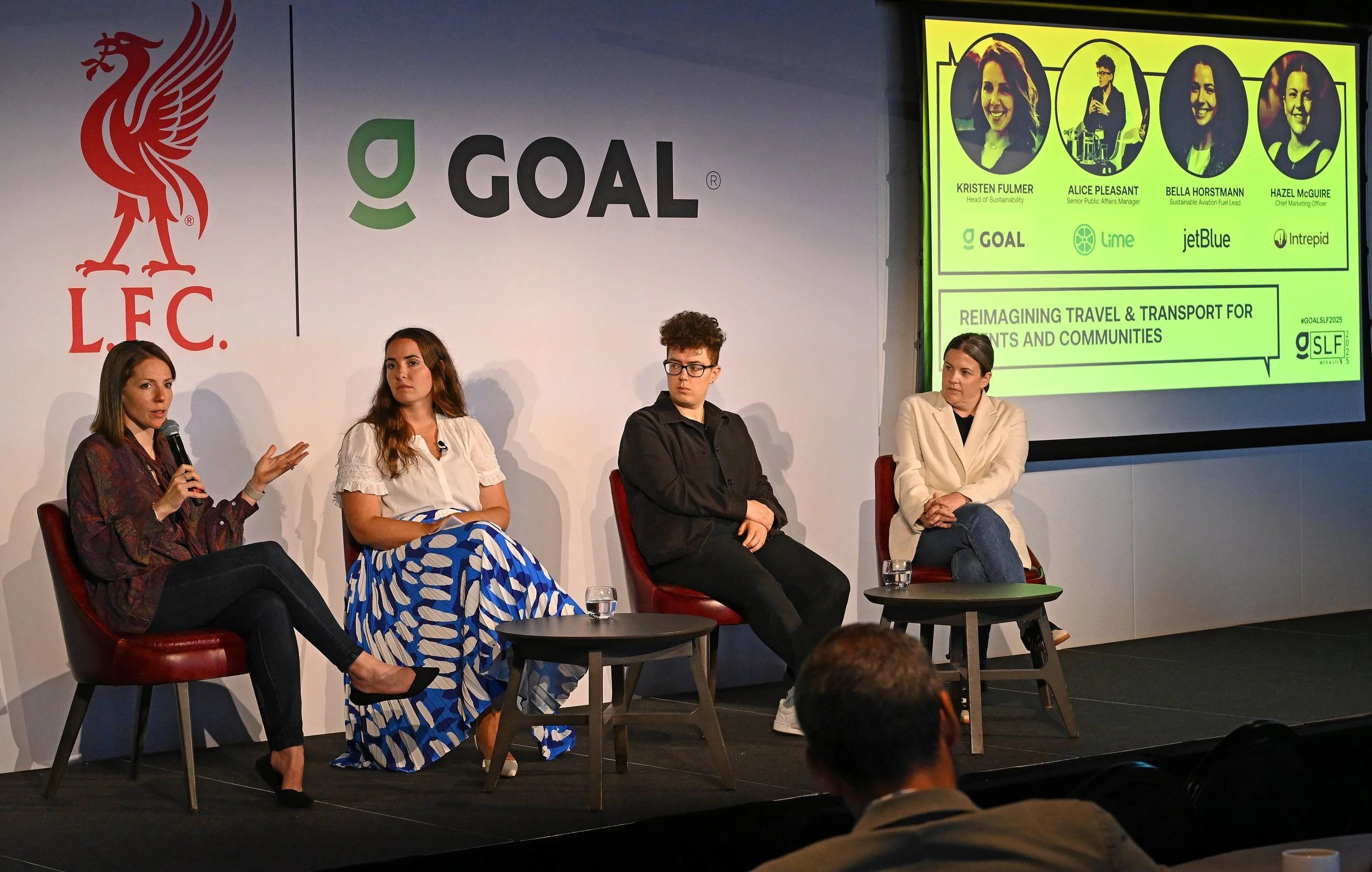 Panel discussion featuring four women seated on stage, one speaking into a microphone. Behind them are logos for Liverpool Football Club, GOAL, Lime, jetBlue, and Intrepid, with a presentation slide highlighting their names and roles, and discussing reimagining travel and transport for residents and communities.