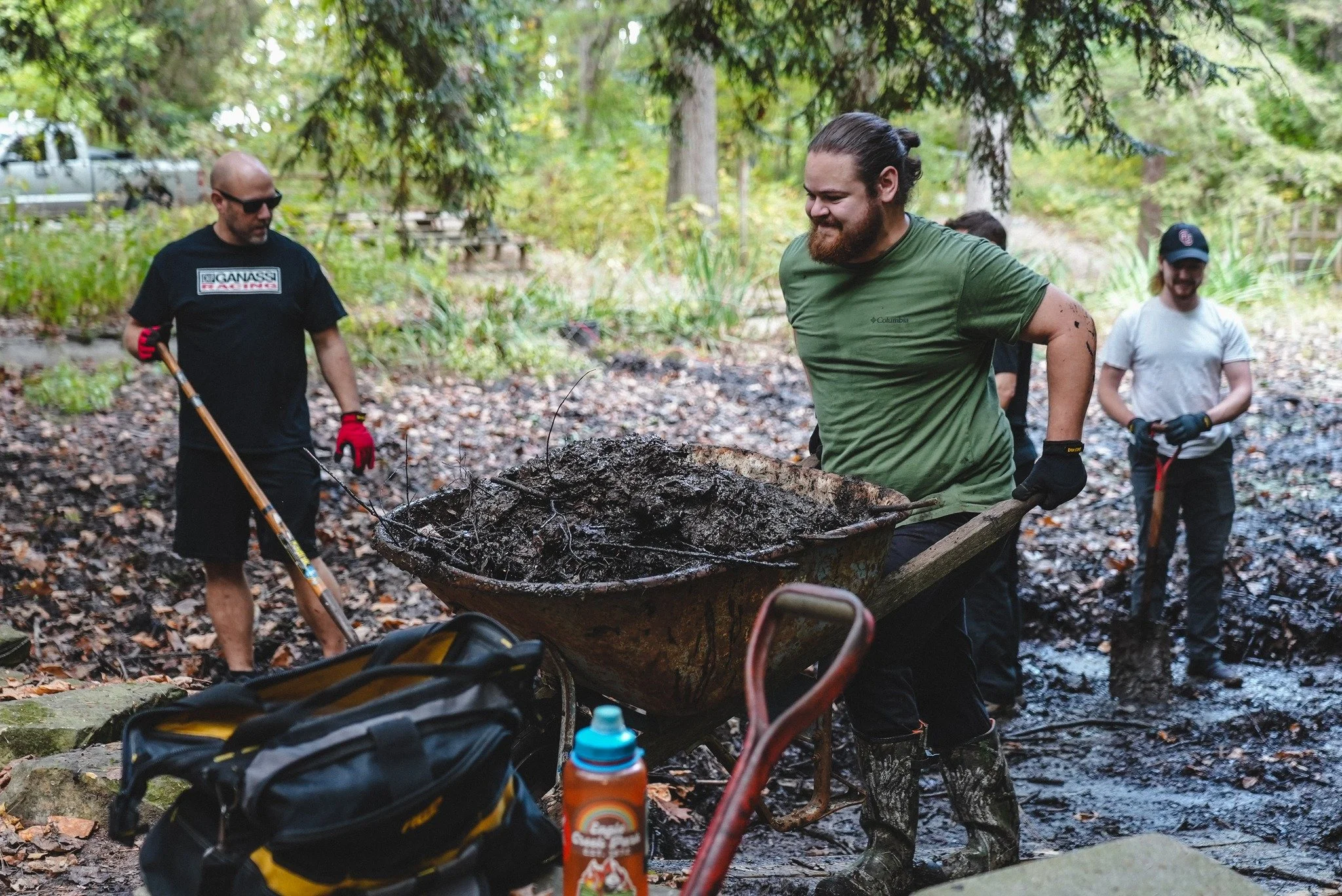 Group of people working together outdoors in a wooded area, with one person pushing a wheelbarrow filled with dirt.