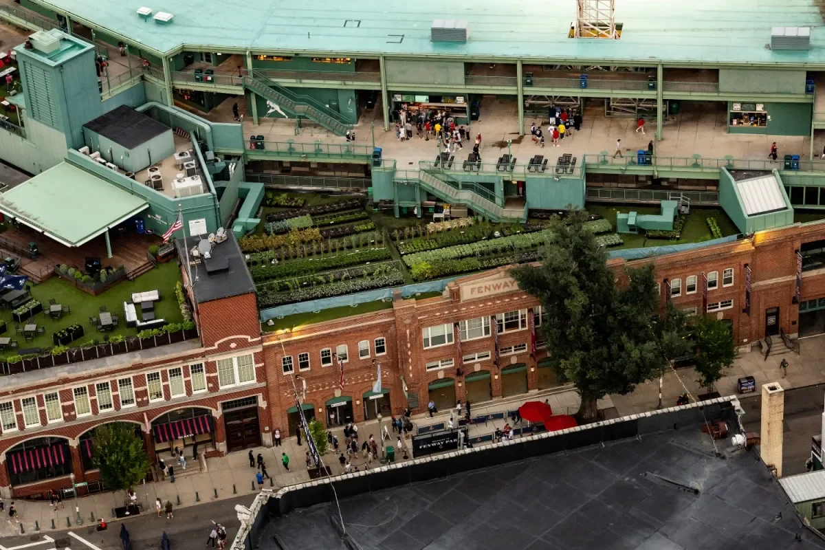 Aerial view of a city street showing a brick building with green rooftop garden and the Fenway Park sign, alongside a large parking area, with pedestrians walking on the sidewalk.