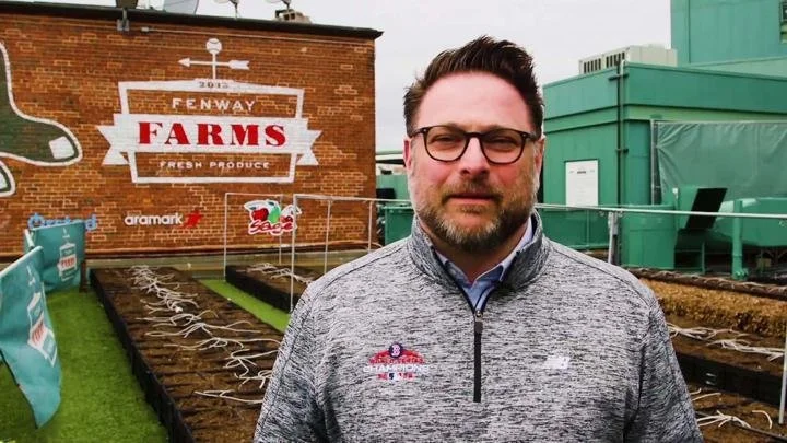 A man with glasses and a beard standing outdoors on a rooftop farm, with vegetable beds and a brick wall with a sign that says "Farrow Farms" in the background.