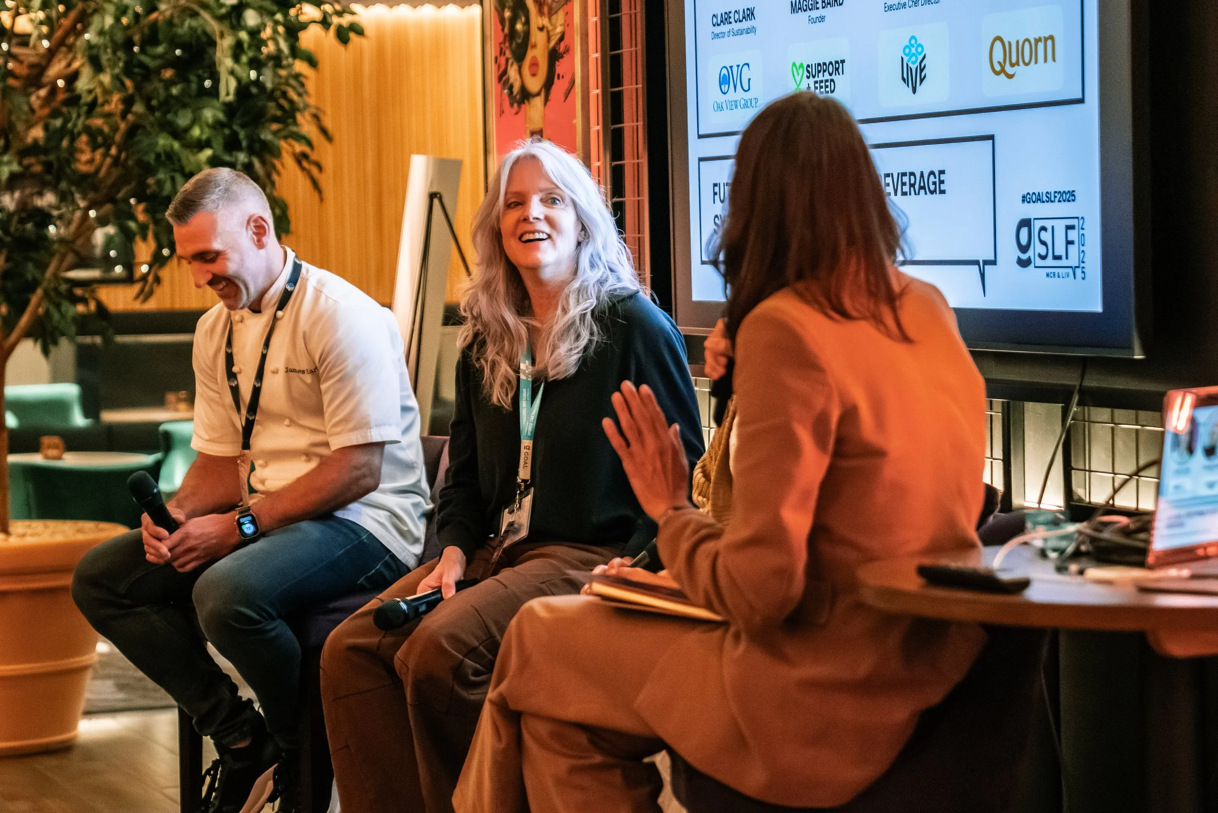 Three people sit on a stage, two women and one man, engaged in a panel discussion at a conference. The woman in the center has long gray hair and is smiling, the woman on the right is speaking into a microphone while facing away from the camera, and the man on the left is holding a microphone and smiling. A screen behind them displays logos and text related to the event.