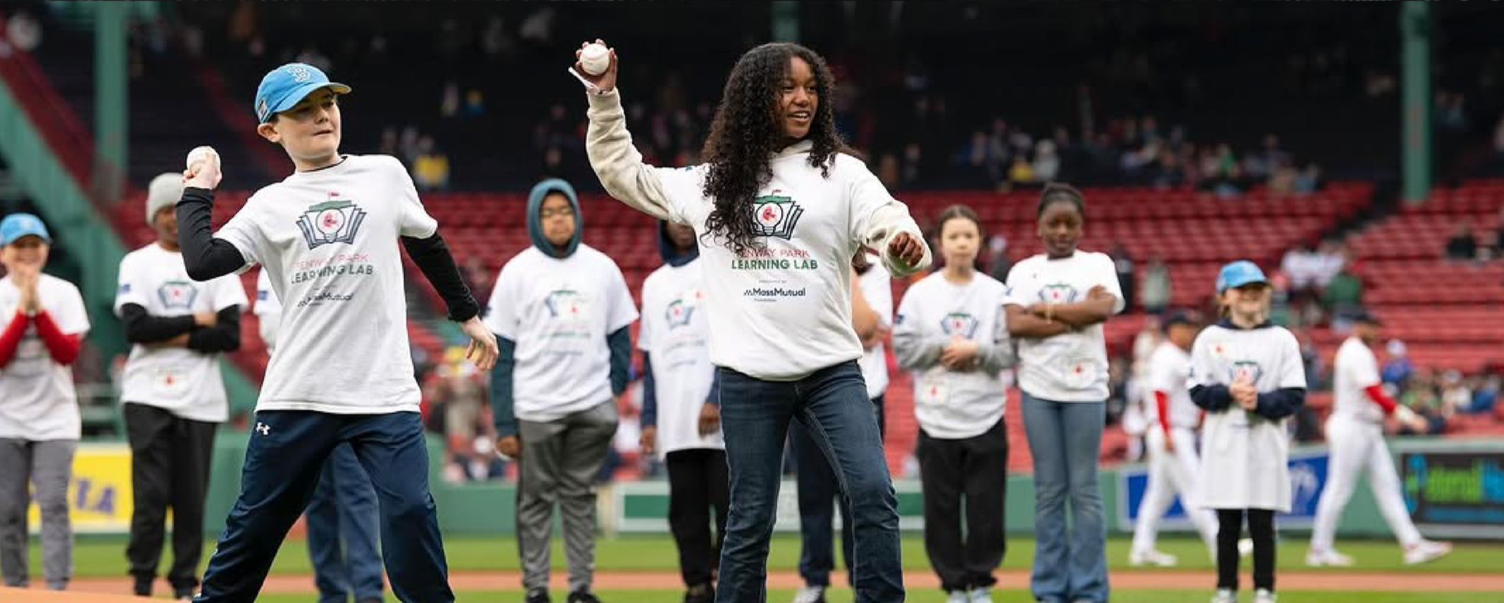 Children and an adult participating in a baseball game at the stadium, wearing white 'Learning Lab' t-shirts. The child in the foreground is holding a baseball, and others are standing on the field with a stadium crowd in the background.