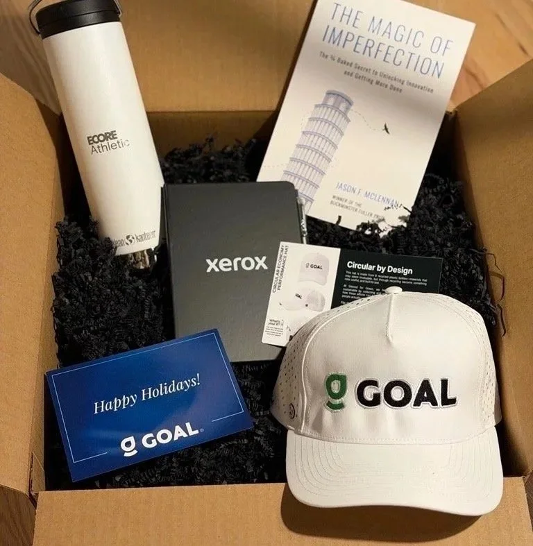 A cardboard box filled with black shredded paper containing a white insulated tumbler, a Bible titled "The Magic of Imperfection," a Xerox notebook, a white GOAL cap, a GOAL greeting card, and A GOAL branded business card.