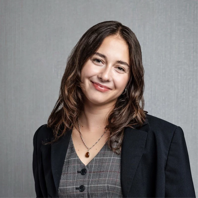 A woman with shoulder-length brown hair, wearing a black blazer over a gray checkered top, smiling softly, standing against a gray background.