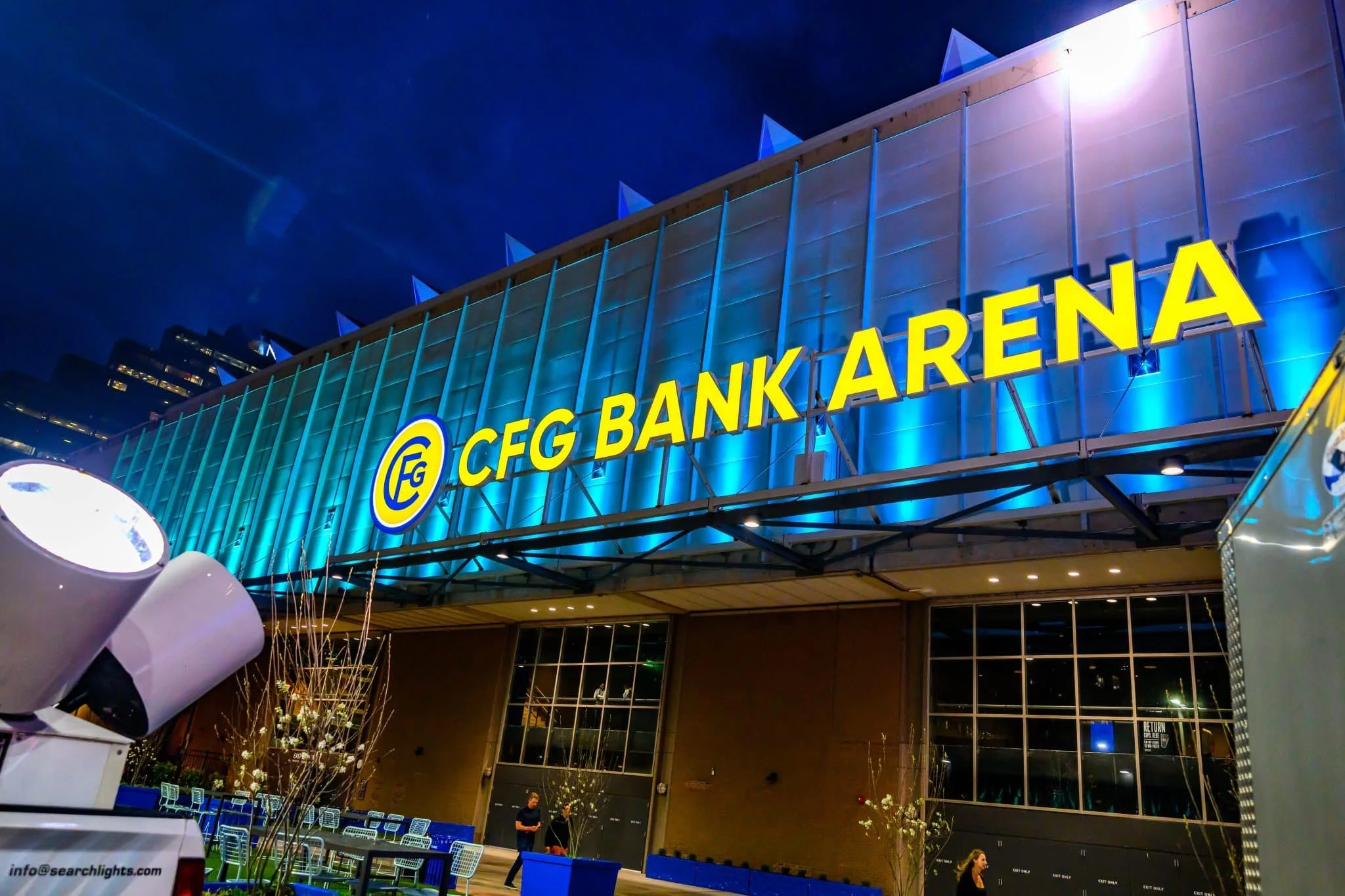Night view of CFG Bank Arena with illuminated blue exterior and yellow sign, with people and seating outside.